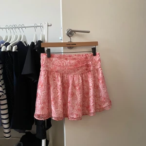 Rosa/orange  mönstrad kortkjol  - Supersöt rosa kortkjol med vitt paisleymönster. Kjolen har bred resår i midjan på baksidan och ett lätt, flowy tyg som ger en skön och avslappnad vibe. Perfekt för varma dagar när du vill känna dig både chill och stylish. Endast använd 3 gånger.