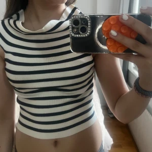 Randig croppad topp i stickad bomull - Säljer en croppad stickad topp med svarta och vita ränder. Toppen kan bäras både offshoulder och med en bredare krage. Den är ärmlös och har en tight passform som framhäver midjan. Perfekt för dig som gillar en trendig och stilren look.