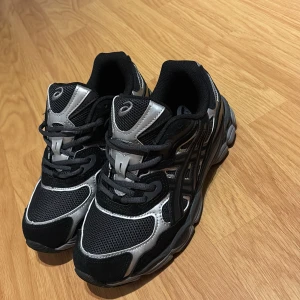 Svart/gråa Asics  - Snygga Asics  i svart och grå med meshpaneler och detaljer i syntet. Skorna har rund tå, platt sula och snörning. Perfekta för dig som gillar sportig stil och vill ha bekväma skor med cool design. (Hör av dig vid minsta fundering)