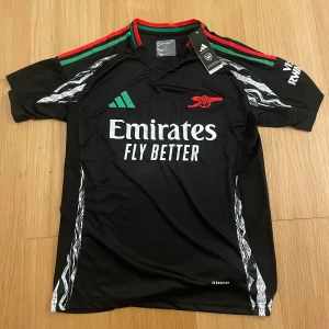 Arsenal svart Adidas fotbollströja M - Svart Arsenal fotbollströja från Adidas med gröna och röda detaljer på axlarna. Tryck med Emirates Fly Better på bröstet och klubbens logga i rött. Tillverkad i lätt funktionsmaterial (AEROREADY) och har korta ärmar samt vita grafiska mönster längs sidorna.