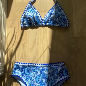 Blåvit blommig bikini - Snygg bikini med blått och vitt blommönster och triangelformad överdel. Bikinin har dekorativa blå kanter. Perfekt för stranden eller poolen.