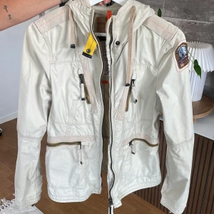 Parajumpers höstjacka - Snygg beige vindjacka från Parajumpers med luva, dragkedjor och små detaljer. Perfekt nu till hösten med en stilren och funktionell jacka till vardags. Storlek S