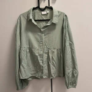 Ljusgrön skjorta från Vila, strl 38 - Superfin ljusgrön skjorta från Vila i storlek 38. Skjortan har knappar framtill, lätt puffade långärmar med resår vid ärmslut och en lös, flowy passform. Tunt bomullstyg med diskret randig struktur. Perfekt för vår och sommar!