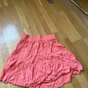 Korallrosa volangkjol i bomull  - Superfin korallrosa kortkjol med bred resår i midjan och volangdetaljer. Kjolen är luftig och har en lätt skrynklig look, perfekt för varma dagar. Tillverkad i mjuk bomull som känns skön mot huden.