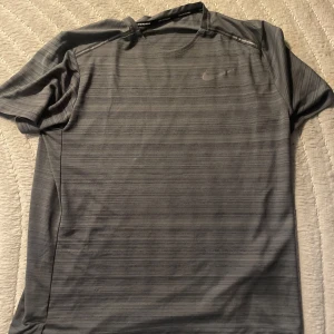 Grå randig Nike t-shirt - Snygg grå t-shirt från Nike med diskreta horisontella ränder och klassisk Nike-logga på bröstet. T-shirten har korta ärmar och är gjord i ett lätt och funktionellt material som passar perfekt för träning eller en aktiv vardag.