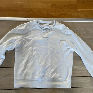 Vit Calvin Klein sweatshirt - Snygg vit sweatshirt från Calvin Klein Jeans med logga på bröstet och tryckt text runt halsringningen. Klassisk passform och långa ärmar, perfekt för en clean och stilren look. Tillverkad i mjuk bomull för extra komfort.