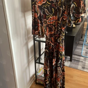 Långklänning med färgstarkt blommönster - Snygg långklänning med svart botten och färgstarkt blommönster i orange, blått, gult och beige. Klänningen har trekvartsärmar och ett löst, flowigt fall. Perfekt för dig som gillar statement-plagg och bohemisk stil.