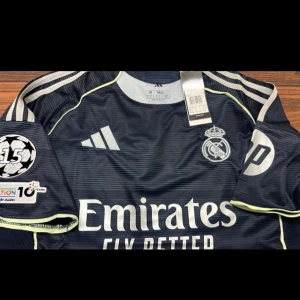 Real Madrid Mbappé borta tröja 2025-26 - Snygg Real Madrid fotbollströja i mörkblått med vita detaljer och tunna horisontella ränder. klubbmärke och sponsortryck på bröstet. Nummer 10 och Mbappé på ryggen. Tillverkad i lätt och ventilerande material, perfekt för match eller träning. Helt ny och kommer med taggar