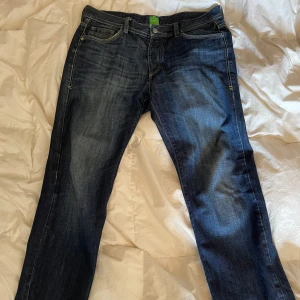 Blå jeans från BOSS Hugo Boss - Snygga blå jeans från BOSS Hugo Boss med klassisk femficksdesign och coola slitningar. Jeansen har raka ben och gröna detaljer på etiketten och fickorna. Perfekta för dig som gillar en stilren men ändå avslappnad look. W 33 L 32. Kom gärna med prisförslag! 