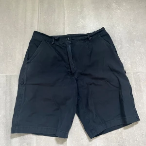 Svarta bomullsshorts med fickor - Svarta shorts i bomull med klassisk passform och raka ben. De har två sidofickor fram och två bakfickor med lock. Midjan är elastisk baktill för extra komfort. Perfekta för varma dagar och enkel att matcha med det mesta.