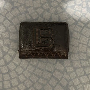 Brun plånbok från LB i skinn - Snygg brun plånbok från LB i glansigt skinn med präglad logga på framsidan. Plånboken har flera fack för kort, mynt och sedlar samt dragkedja och tryckknapp. Perfekt för dig som vill ha ordning på allt i en stilren accessoar.