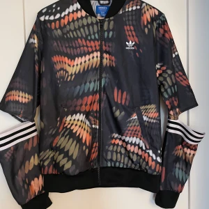 Adidas träningsjacka med färgstarkt mönster designade av Rita Ora - Snygg Adidas träningsjacka med färgstarkt abstrakt mönster i svart, orange, grön och vit. Jackan har dragkedja, två fickor och klassiska vita Adidas-ränder på ärmarna. Stor Adidas-logga på ryggen och mindre logga fram. Perfekt statement-plagg för dig som vill sticka ut.