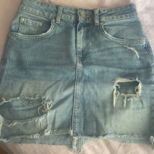 Ljusblå jeanskjol med slitningar - Snygg ljusblå jeanskjol från gina med slitna detaljer och fransig nederkant. Klassisk femficksmodell med bälteshällor och dragkedja fram. Perfekt för dig som gillar en avslappnad och trendig stil. 