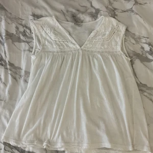 Vit blus med spets från Lucky Brand - Supersöt vit blus från Lucky Brand med v-ringning och broderade spetsdetaljer upptill. Ärmlös modell med lätt rynk under bysten som ger en luftig och avslappnad känsla. Perfekt för varma dagar och enkel att styla med jeans eller shorts.