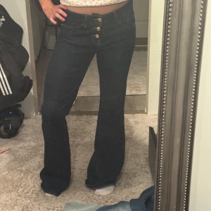 Mörkblå bootcut jeans med knappar - Säljer ett par mörkblå jeans med bootcut passform och medellågmidja. Byxorna har snygga guldiga knappar framtill och klassiska fickor både fram och bak. Jeansen är i stretchigt denimtyg och har kontrastsömmar för en extra detalj.