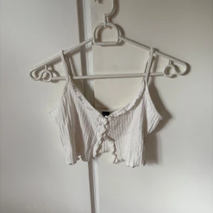 Vit ribbad croppad topp från SHEIN - Supersöt vit croppad linnetopp från SHEIN med smala axelband och ribbad struktur. Toppen har knappar framtill och en lätt vågig kant nertill som ger en extra detalj. Perfekt för varma dagar eller att styla lager på lager.