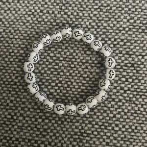 Vitt pärlarmband med svarta kors - Coolt armband med vita runda pärlor och svarta kors på varje pärla. Stretchig modell som passar de flesta handleder. Perfekt accessoar för dig som gillar unika detaljer och vill sticka ut lite extra. Passar yngre barn 