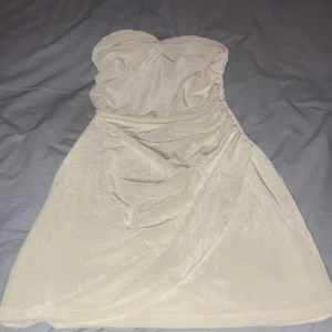 Glittrig vit axelbandslös klänning BikBok - Supersnygg axelbandslös kortklänning från BikBok i glittrigt vitt tyg. Klänningen har en omlottliknande drapering framtill och är figurnära med en mjuk, stretchig känsla. Perfekt för dig som vill sticka ut med en elegant och trendig look.