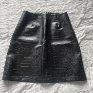 Svart kjol i krokodilskinn - Snygg svart kjol i fejkskinn med krokodilmönster och rak passform. Dragkedja bak för enkel påklädning. Perfekt för dig som vill sticka ut lite extra. Går att klä upp och klä ner.