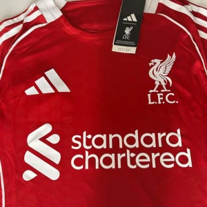 Liverpool FC 25/26 hemmatröja - Liverpool FCs officiella matchtröja från Adidas i rött med vita detaljer och klubbmärke på bröstet. Tröjan har klassiska Adidas-ränder på axlarna och sponsorlogga framtill. Tillverkad i lätt och ventilerande funktionsmaterial.