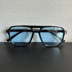 Svarta solglasögon med blåtonade glas - Stilrena pilotglasögon med svart ram och blåtonade glas. Bågarna har en klassisk aviator-form med dubbla nitar vid sidorna och en markerad brygga.💯Perfekta för dig som vill ha en cool och retro vibe till din look. Hör av dig vid minsta fundering!🤩