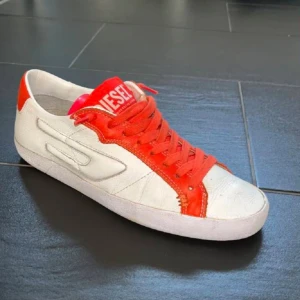 Vita och orange sneakers från Diesel - Snygga sneakers från Diesel i vitt skinn med röda detaljer och snörning. Klassisk låg modell med rund tå och platt sula. Perfekta för dig som vill sticka ut med en sportig och färgstark look.