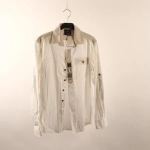 Vit skjorta från Haus Huberts - Vit långärmad skjorta från Haus Huberts med beige axelparti och krage. Snygga detaljer längs knappslån och en bröstficka med knapp. Skjortan har klassisk passform och knappar framtill. Perfekt för dig som gillar traditionell stil med en twist.