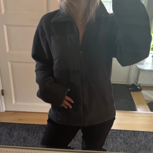 Svart teddytröja från McKinley - Mysig svart teddytröja/tunnare jacka från McKinley som skiftar i grå gröna toner. Med hel dragkedja och en praktisk bröstficka. Jackan har en fin krage och långa ärmar. Perfekt när du vill vara både varm och snygg.