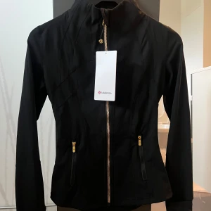 Svart träningsjacka från Lululemon - Snygg svart träningsjacka från Lululemon med guldiga dragkedjor och figurnära passform. Jackan har hög krage, långa ärmar och två fickor med dragkedja framtill. Perfekt för dig som vill ha en sportig och stilren look.