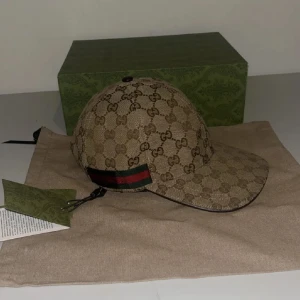 Gucci keps med GG-mönster, beige  - Exklusiv keps från Gucci i beige canvas med klassiskt GG-monogram över hela kepsen. Har en grön och röd rand på sidan och justerbar rem baktill i brunt läder. Insidan är fodrad i mörkbrunt tyg. Perfekt för dig som vill sticka ut med lyxig streetstyle.