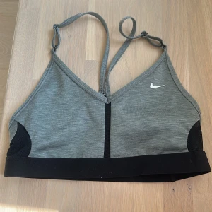 Nike grå sporttopp Dri-FIT S - Sportig grå topp från Nike med svarta detaljer och justerbara tunna axelband. Tillverkad i Dri-FIT-material som andas och håller dig torr under träningen. Snygg V-ringning och bred svart resår nedtill för extra stöd. Perfekt för gymmet eller löprundan.