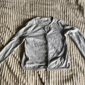 Ljusgrå kofta från H&M med knappar - En stilren ljusgrå kofta från H&M med rund halsringning och knäppning framtill. Tröjan har långa ärmar och ribbade muddar vid ärmslut och nederkant. Perfekt att slänga över en t-shirt för en chill look. Kommer från ett hem med katt.