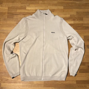 Beige kofta med dragkedja från BOSS - Snygg beige kofta från BOSS med hel dragkedja framtill och diskret logga på bröstet. Tillverkad i stretchig bomull med ventilerande mesh-detaljer vid axlar och rygg. Perfekt för lager-på-lager och stilren look. Denna är aldrig anvönd