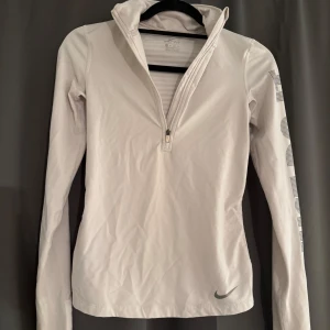 Vit Nike Dri-Fit träningströja half zip - Superskön vit långärmad träningströja från Nike med Dri-Fit material. Tröjan har half zip framtill och trycket 'JUST DO IT' i grått längs ena ärmen. Perfekt för löpning eller gym och sitter snyggt med en slim passform.