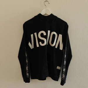 Woxo hoodie - Svart hoodie från Woxo med stor vit VISION-tryck på ryggen och textdetaljer längs ärmarna. Tröjan har huva och är tillverkad i mjuk bomullsblandning. Perfekt för dig som gillar streetwear och vill sticka ut med en cool look.