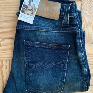 Mörkblå jeans från Nudie Jeans - Snygga mörkblå jeans från Nudie Jeans med klassisk femficksdesign och coola slitningar. Jeansen har kontrastsömmar och en läderpatch med logga bak i midjan. Perfekta för dig som gillar en avslappnad men stilren look.