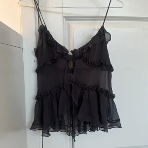 Svart volangig blus från Zara XS - Svart blus från Zara i transparent, lätt material. Topp med tunna axelband, volanger och knytning framtill. Använd fåtal gånger och är i fint skick. Har förkortat axelbanden själv men det går enkelt att sprätta/klippa upp om man önskar att ha axelbanden längre. 