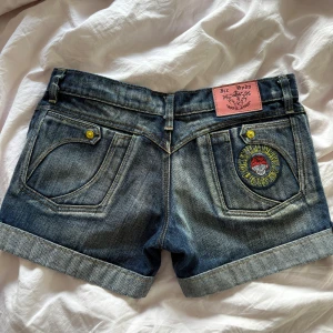 Jeansshorts från Ice Soda med brodyr - Snygga jeansshorts från Ice Soda i mörkblå denim med uppvikta benslut, coola spänndetaljer och broderad patch på bakfickan. Gula knappar och unika sömmar ger extra edge. Perfekta för en trendig sommarlook! PRIS KAN DISKUTERAS 