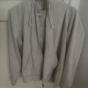 Vit hoodie  med dragkedja H&M - Enkel och stilren vit hoodie från H&M i storlek M. Tröjan har huva med snörning, dragkedja framtill och två fickor. Perfekt för chill dagar eller när du vill ha en avslappnad look.