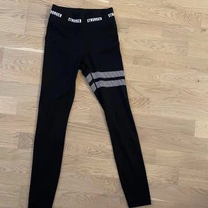 Svarta leggings från Stronger  - Snygga svarta leggings från Stronger i storlek XS. Hög midja med logga runtom och två vita randiga detaljer på ena benet. Tillverkade i stretchigt material med 73% polyester och 27% elastan – perfekta för träning eller chill. Aldrig använda!