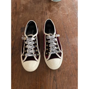 Sneakers från Christian Dior - Snygga sneakers från Christian Dior i bordeaux sammet med vita detaljer och gummisula. Skorna har vita snören med Christian Dior-tryck. Klassisk rund tå och låg modell, perfekta för dig som vill ha ett par coola sneakers som livar upp vilken outfit som helst! 