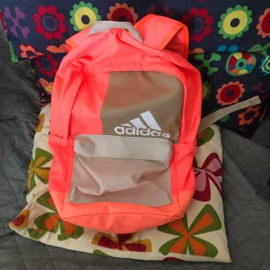 Neonorange Adidas ryggsäck - Säljer en snygg neonorange ryggsäck från Adidas med beige detaljer och vit logga framtill. Väskan har ett stort huvudfack, en praktisk ytterficka och justerbara axelremmar. Perfekt för skolan eller träningen när du vill sticka ut lite extra.