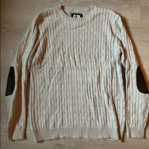 Beige kabelstickad tröja Made in the Shade - Kabelstickad beige tröja från Made in the Shade i storlek L. Tröjan har rund hals, ribbade muddar och mörka mockadetaljer på armbågarna. Perfekt för dig som gillar klassisk stil. 