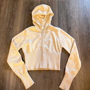 Beige croppad hoodie från Gina Tricot - Beige croppad hoodie från Gina Tricot med dragkedja framtill och huva. Tröjan är stickad med bred ribbad mudd vid midjan och ärmsluten. Använd 2 gånger.🌺