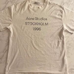Vit Acne Studios t-shirt Stockholm 1996 - Vit t-shirt från Acne Studios med tryck 'Acne Studios STOCKHOLM 1996' på bröstet. Klassisk passform med rund hals och korta ärmar. Tillverkad i 100% bomull för en skön och avslappnad vibe. Pris kan diskuteras 
