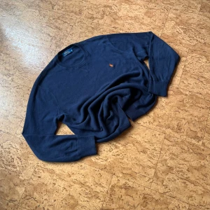 Mörkblå stickad tröja Polo Ralph Lauren - Mörkblå stickad tröja från Polo Ralph Lauren med klassisk rund hals och broderad orange logga på bröstet. Tröjan har långa ärmar och ribbade muddar. Perfekt för dig som gillar stilren och tidlös design.