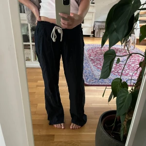 Svarta linnebyxor från Roxy med snörning - Svarta linnebyxor från Roxy med avslappnad passform och breda ben. Byxorna har resår i midjan och vit snörning framtill. Rå kant nertill för en chill vibe. Perfekta för dig som gillar bekväma och snygga byxor till vardags.