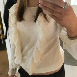 Vit blus med volangdetaljer - Supersöt vit blus med diskret prickigt mönster och stora volanger framtill. Långärmad modell med lätt transparent tyg som ger en luftig känsla. Perfekt att styla med jeans eller kjol för en trendig look. I strl 164 men passar större strl med 