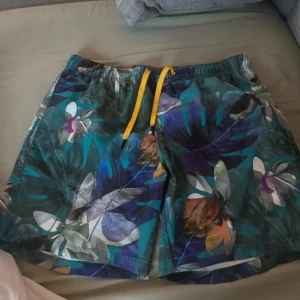 Blommiga badshorts från Björn Borg - Färgglada badshorts från Björn Borg med blommigt mönster i blått, lila, grönt och inslag av vitt och brunt. Shortsen har gul snörning i midjan och meshfoder på insidan. Perfekta för stranden eller poolen i sommar.
