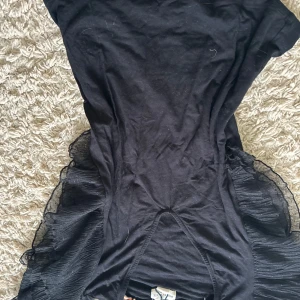 Svart tröja Holly & White - Svart tröja från Holly & White med coola volangdetaljer i mesh på sidorna. Klänningen har en enkel, figurnära passform och är tillverkad i mjukt bomullstyg. Perfekt för dig som gillar en stilren look med en twist.💕